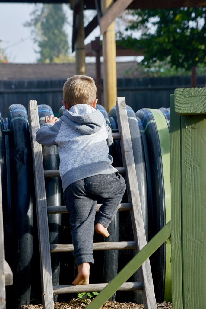 Young child climbing barefoot up a wooden ladder onto a large outdoor play structure at a nature-based school.