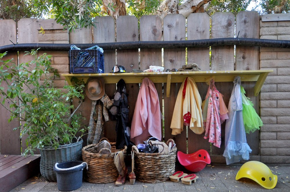 Outdoor preschool dress-up corner with baskets of costumes, hats, boots, and hanging capes for imaginative play.