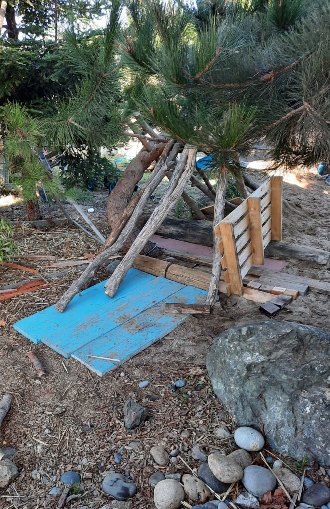 Outdoor child-built fort made from wooden pallets, planks, and tree branches beneath pine trees, encouraging creativity and problem-solving.