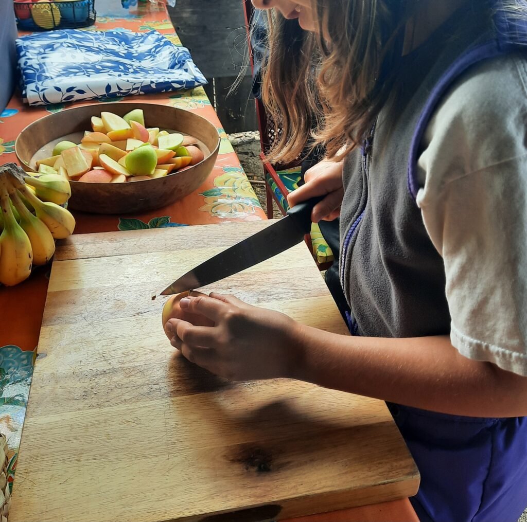 Child slicing apples on a wooden cutting board during a snack preparation activity with fresh fruit.
