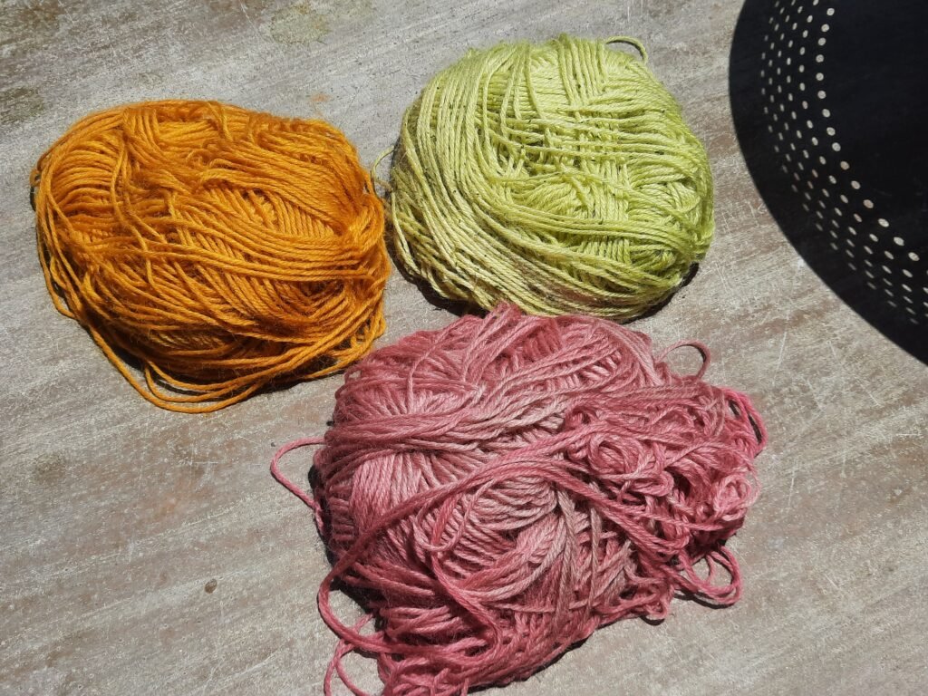 Three balls of naturally dyed yarn in orange, green, and rose pink shades displayed on a wooden surface in sunlight.