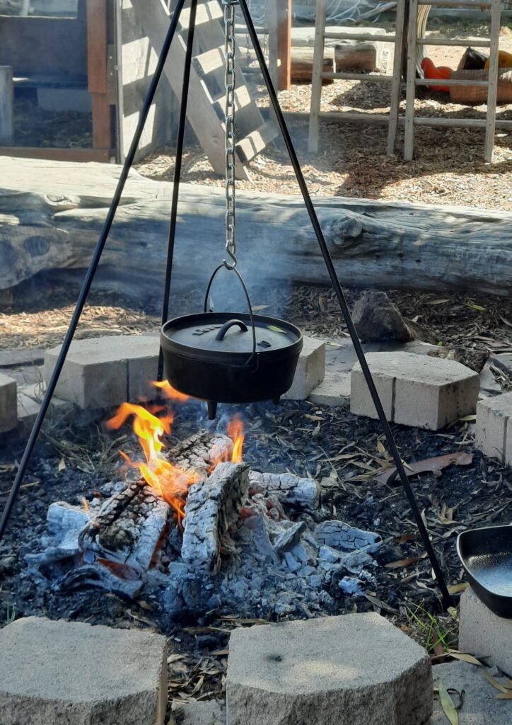 Black cast iron pot with lid hanging from a tripod over a wood fire in an outdoor fire pit area.