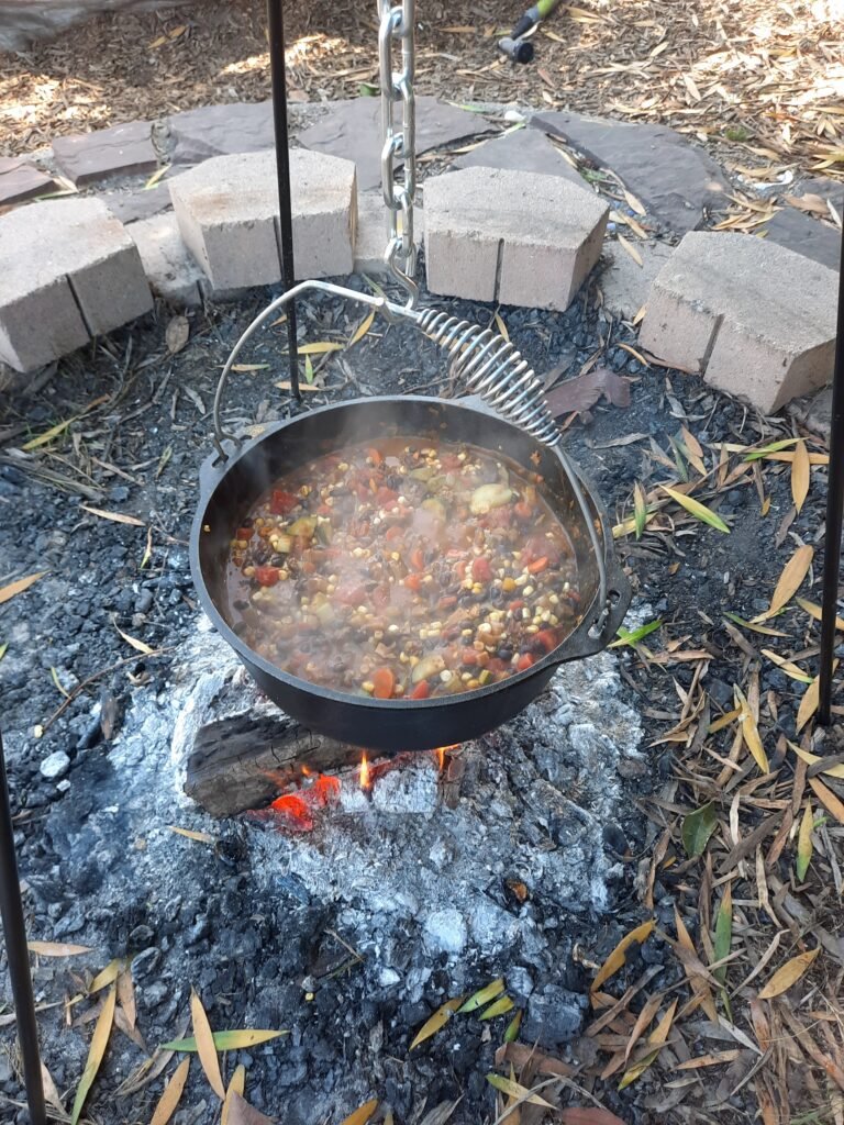 Cast iron pot filled with colorful stew simmering over an open campfire surrounded by bricks and ashes