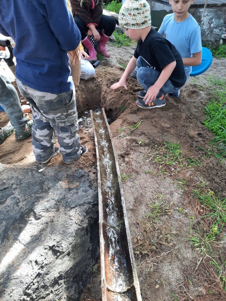 Children playing in a sand area, channeling water through a wooden trough as they dig and explore together.