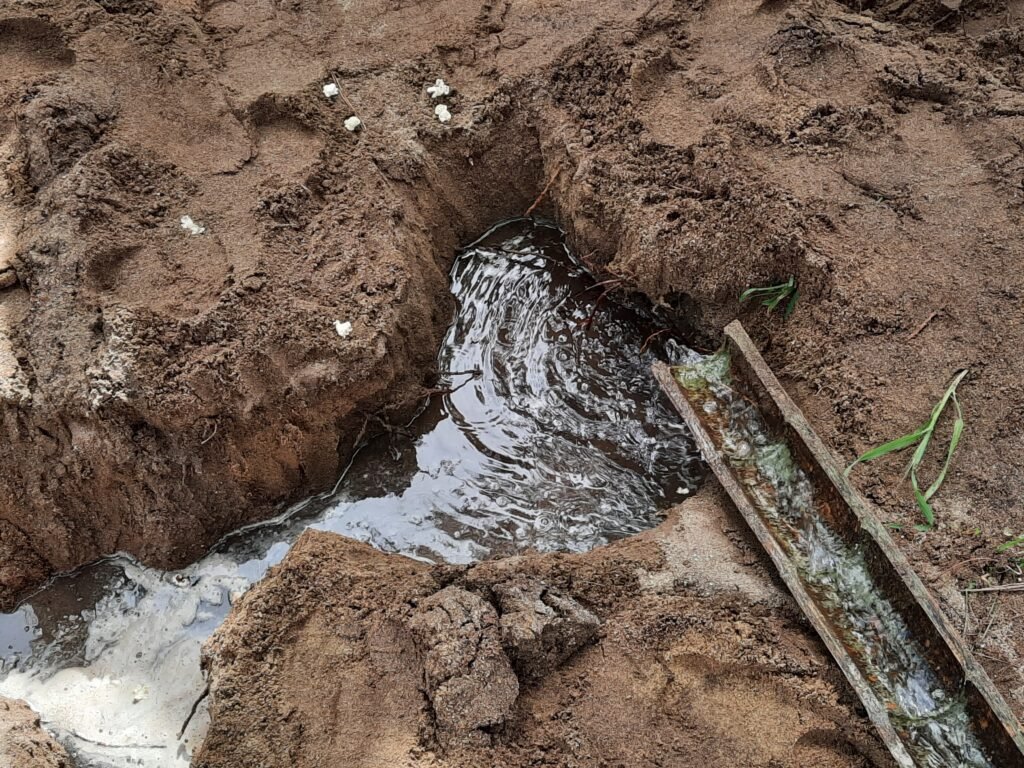 Close-up view of a small water channel carved in dirt, showing flowing water in a wooden trough and small white objects scattered in the sand