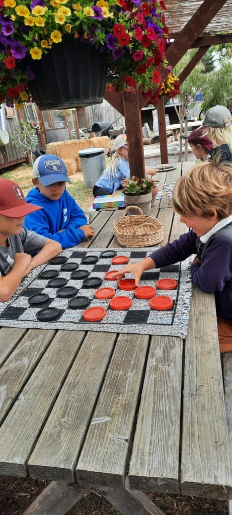 Children sitting at a wooden picnic table playing an oversized checker game with red and black pieces under hanging flower baskets.