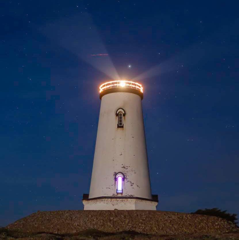 White lighthouse illuminated at night with its beacon shining under a starry sky.