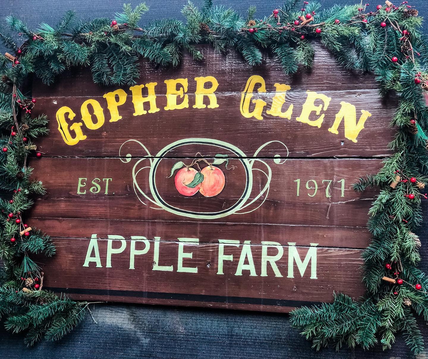 Wooden Gopher Glen Apple Farm sign decorated with evergreen branches and red berries, featuring two painted apples and the text “Gopher Glen Apple Farm, Est. 1971.”