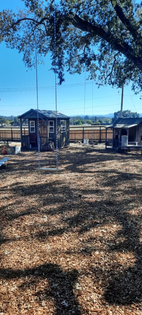 A spacious outdoor play and gathering area covered in wood chips, featuring a firepit surrounded by wooden Adirondack chairs in the center. To the left is a covered wooden pavilion with long picnic tables underneath. To the right stands a small rustic playhouse with a turquoise door, set on a porch decorated with hay bales and small wooden figures. A fenced driveway and parked vehicle are visible in the background, with large trees providing partial shade across the scene.