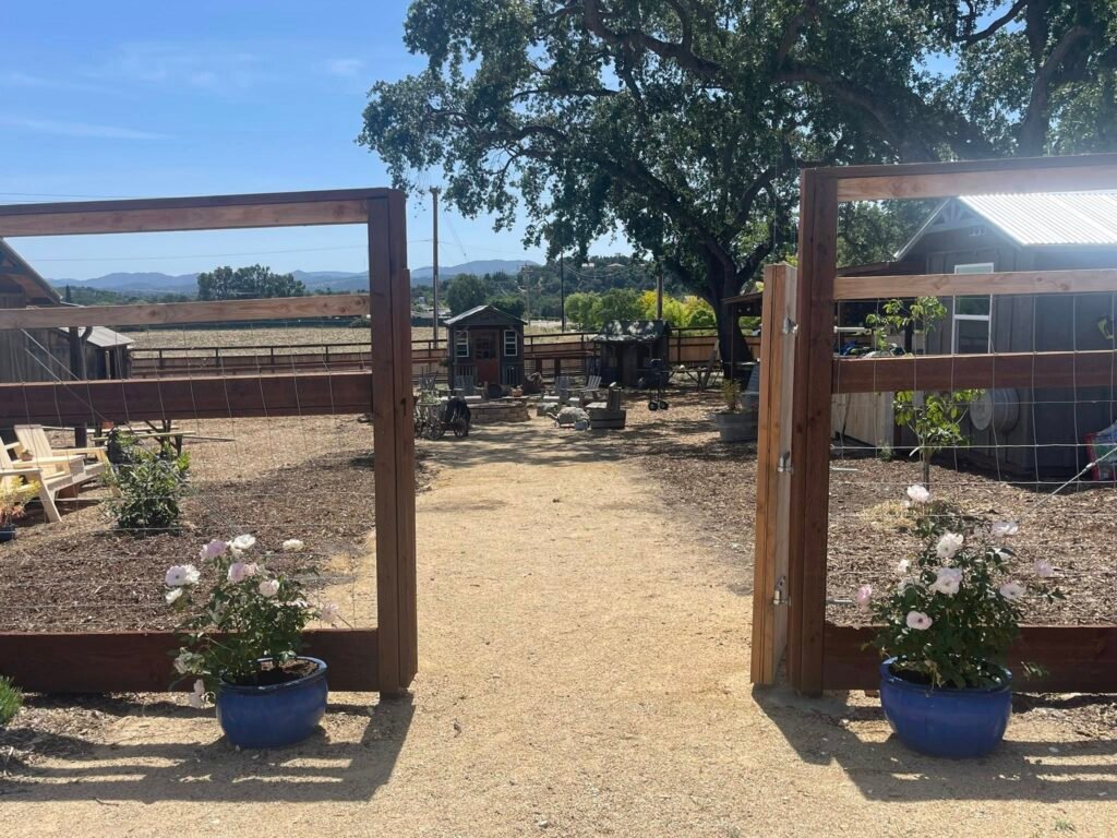 Wide entrance gate leading into a rustic outdoor learning and garden space with gravel pathways, flower pots, wooden fencing, and views of hills in the distance.