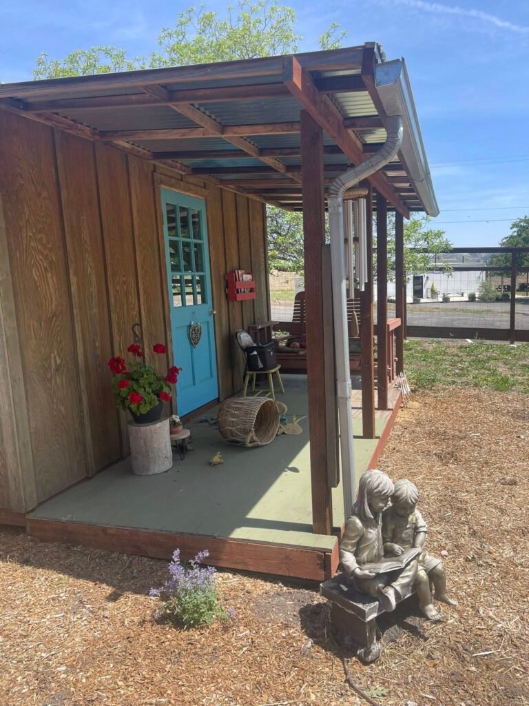 Rustic wooden schoolhouse-style building with a covered porch, bright blue door, hanging wind chimes, red flowers in a planter, and a reading statue of two children in front on a sunny day.