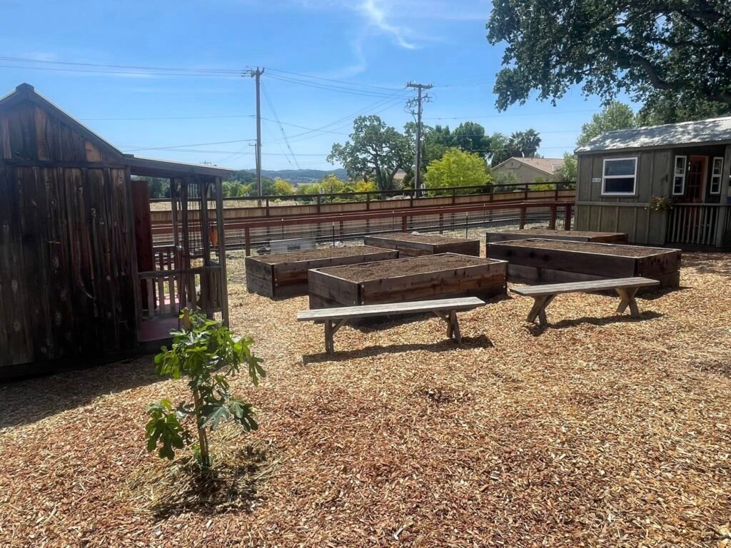 Raised garden beds filled with soil beside rustic wooden buildings and benches, overlooking rolling hills and farmland under a clear blue sky.